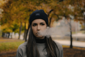 Woman in a black beanie exhaling smoke outdoors against autumn trees during a fall smoke session.